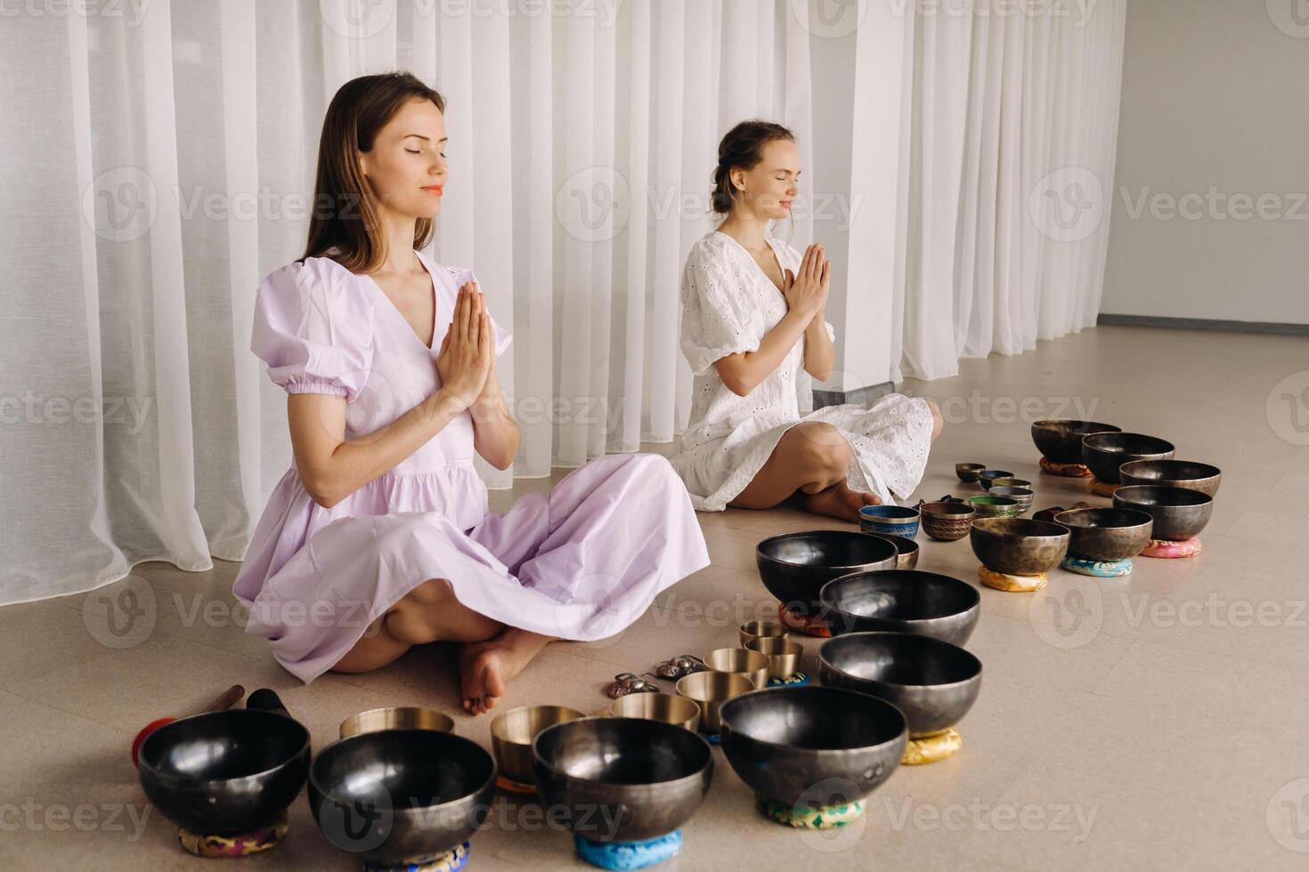 zwei Frauen sind Sitzung mit tibetanisch Schalen im das Lotus Position Vor ein Yoga Klasse im das Fitnessstudio foto