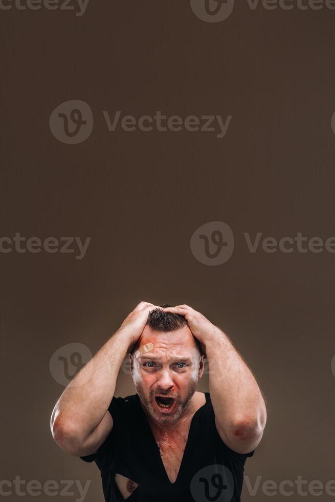 gegen ein grau Hintergrund steht ein angeschlagen wütend Mann im ein schwarz T-Shirt mit Wunden foto