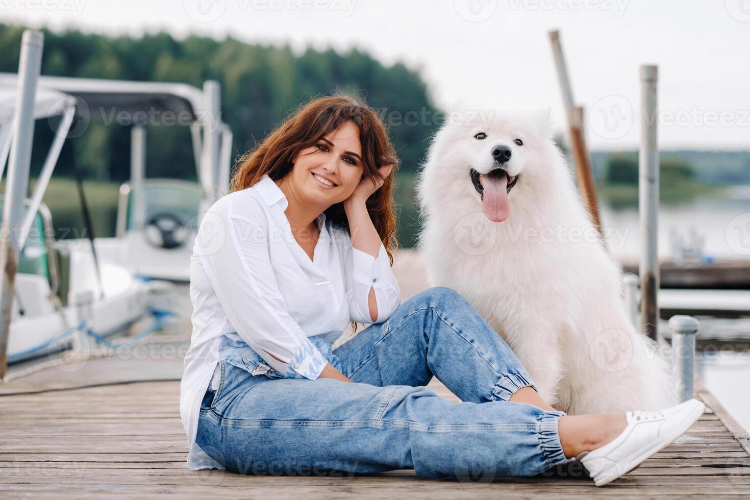 ein glücklich Frau mit ein groß Weiß Hund sitzt auf ein Seebrücke durch das Meer beim Sonnenuntergang foto