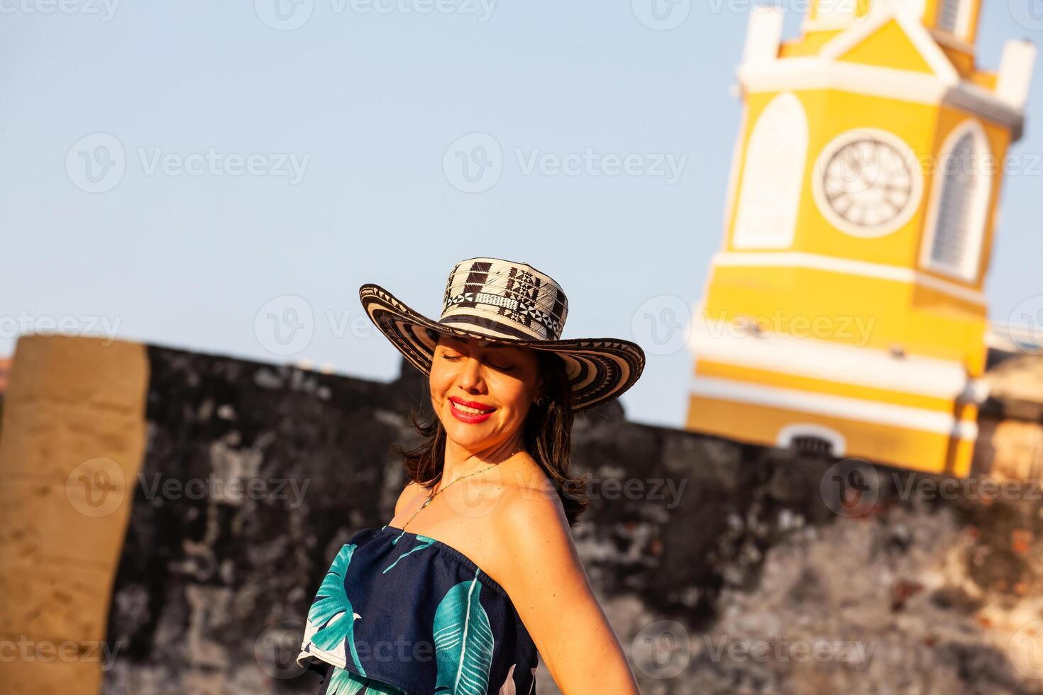 schön Frau tragen das traditionell kolumbianisch Hut namens Sombrero vueltiao beim das Uhr Turm auf das historisch Straßen von das Cartagena de Indien ummauert Stadt foto