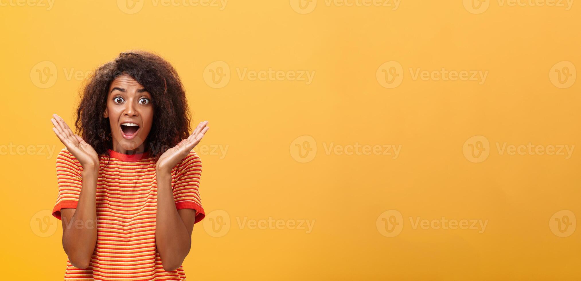 hüfthoch Schuss von erstaunt froh gesellig dunkelhäutig weiblich mit afro Frisur im gestreift T-Shirt gestikulieren mit Palmen angehoben in der Nähe von Gesicht Blick erstaunt und Erfreut beim Kamera von Überraschung und Freude foto