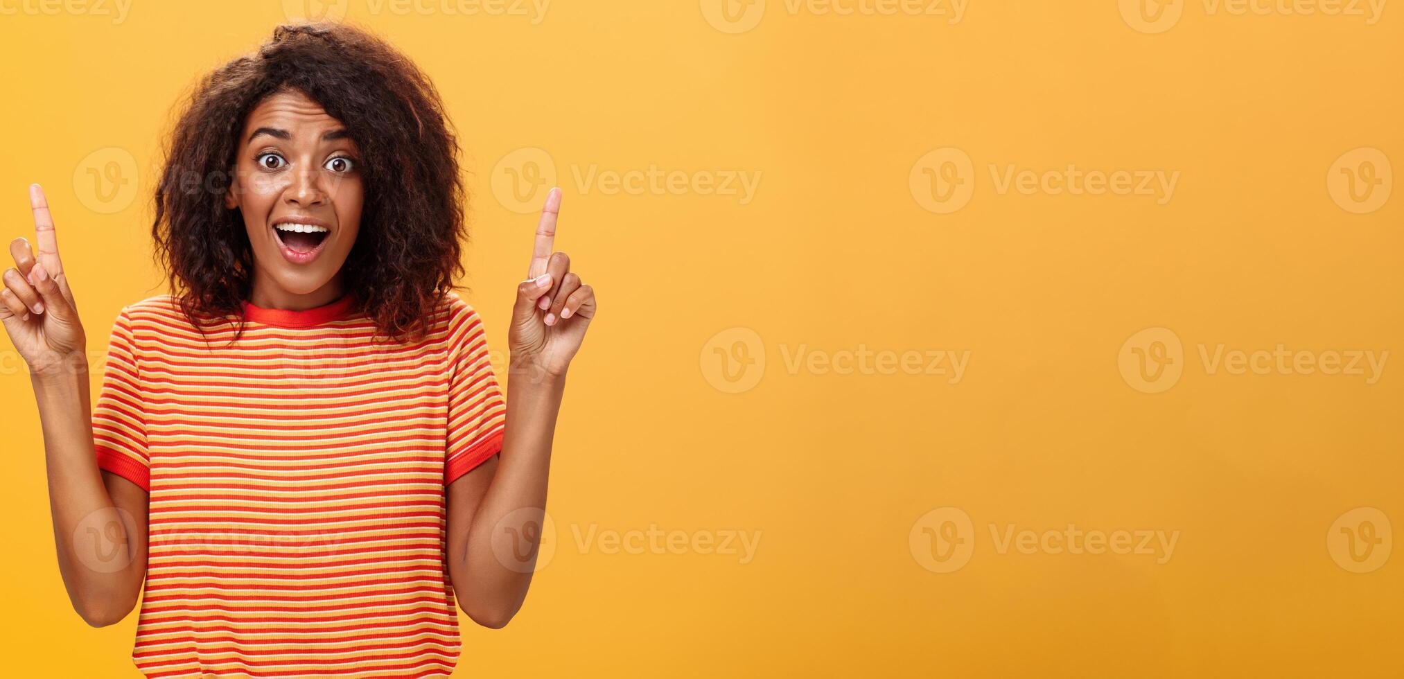 erstaunt glücklich begeistert süß afrikanisch amerikanisch Frau mit lockig Frisur im gestreift T-Shirt erziehen Index Finger zeigen oben Blick fasziniert und Erfreut beim Kamera Über Orange Mauer foto