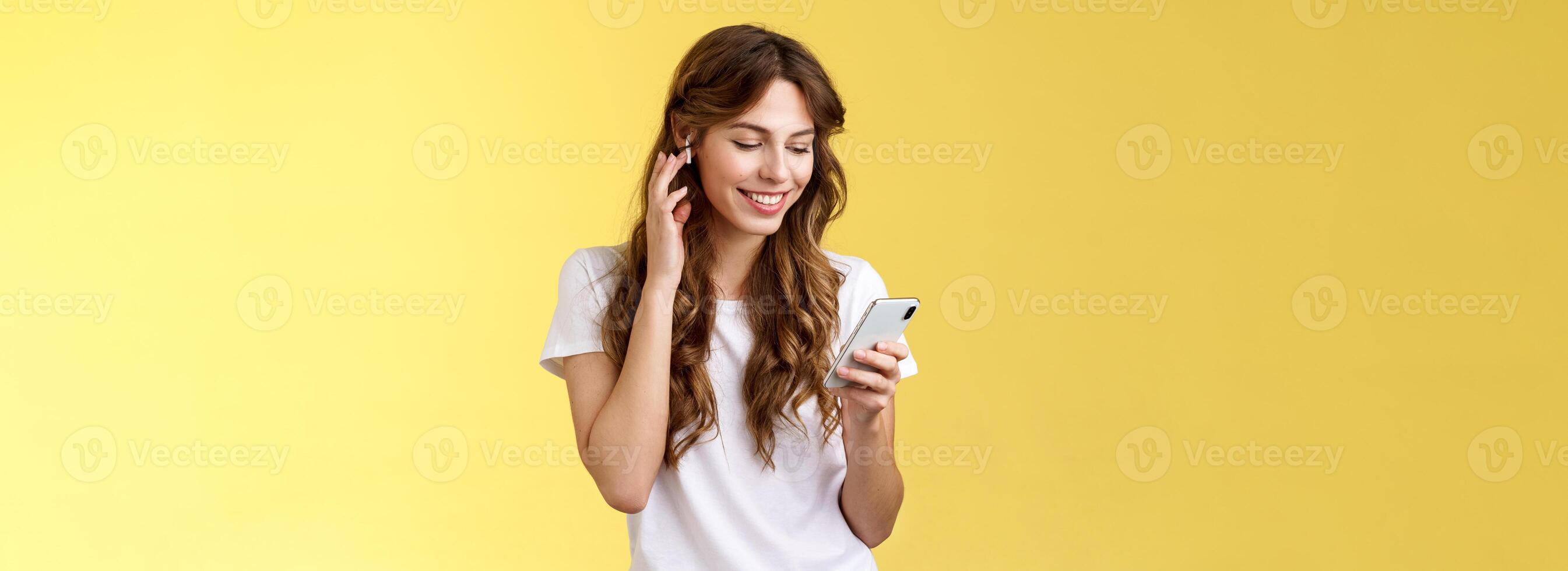 zärtlich feminin lockiges Haar weiblich Weiß T-Shirt berühren kabellos Ohrhörer stellen Kopfhörer Ohr lächelnd zufrieden aussehen Smartphone Bildschirm wählen Lied will Hör mal zu Musik- Suche richtig Spur Gelb Hintergrund foto