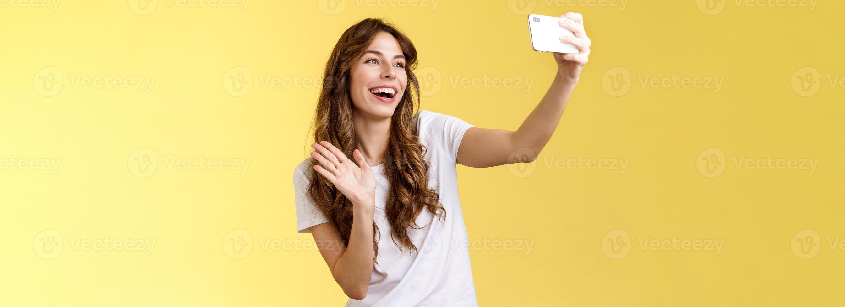freundlich lebhaft gut aussehend heiter feminin Mädchen erweitern Arm halt Smartphone Aufzeichnung Video Blog winken Vorderseite Kamera lächelnd breit reden Fans Videoanruf nehmen Selfie Gelb Hintergrund foto