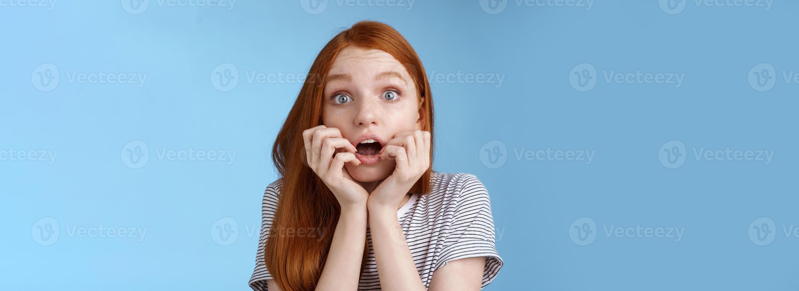 schockiert sprachlos keuchend jung Rothaarige Mädchen starren beeindruckt betäubt Aufpassen wichtig Moment Fernseher Serie beißen Finger öffnen Mund zitterte Stehen aufgeregt Blau Hintergrund vorwegnehmen foto