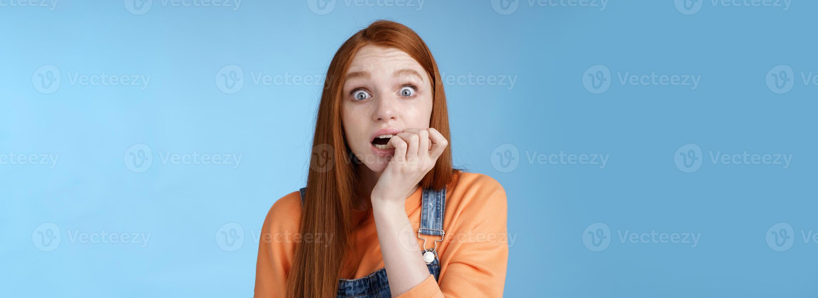 erschrocken unsicher Ängstlich jung Zittern Rothaarige Mädchen breit Augen starren heftig emotional beißen Fingernägel, Ventilator Sorge Liebling Charakter Fernseher Serie stirbt Stehen nervös Blau Hintergrund foto