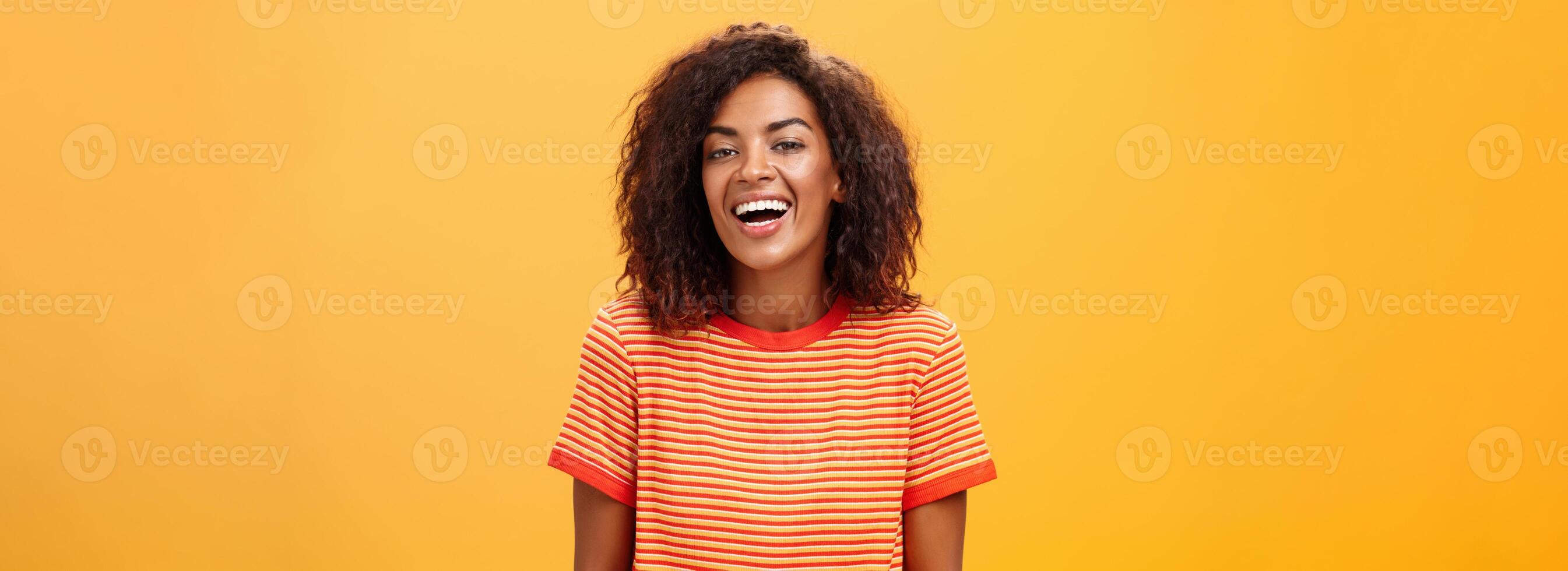 hüfthoch Schuss von abgehend glücklich charmant dunkelhäutig weiblich mit lockig Frisur Lachen freudig posieren im gestreift modisch T-Shirt Über Orange Hintergrund genießen nett beiläufig Konversation foto