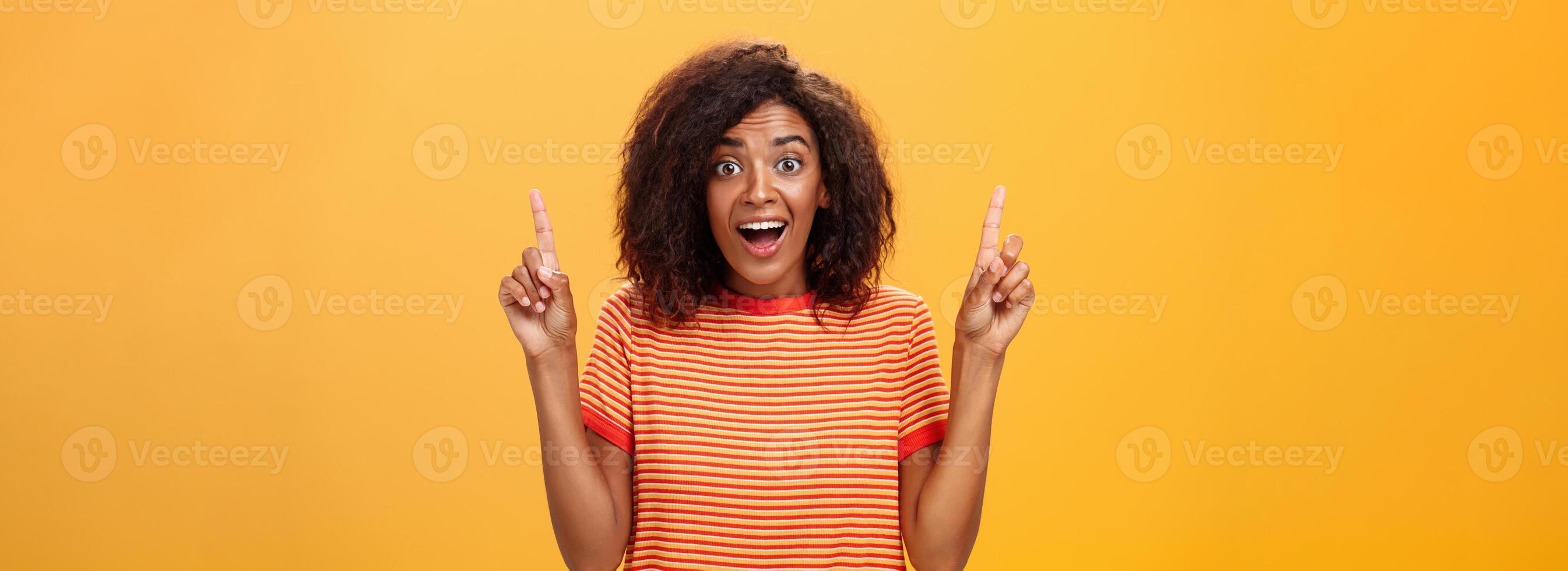 erstaunt glücklich begeistert süß afrikanisch amerikanisch Frau mit lockig Frisur im gestreift T-Shirt erziehen Index Finger zeigen oben Blick fasziniert und Erfreut beim Kamera Über Orange Mauer foto