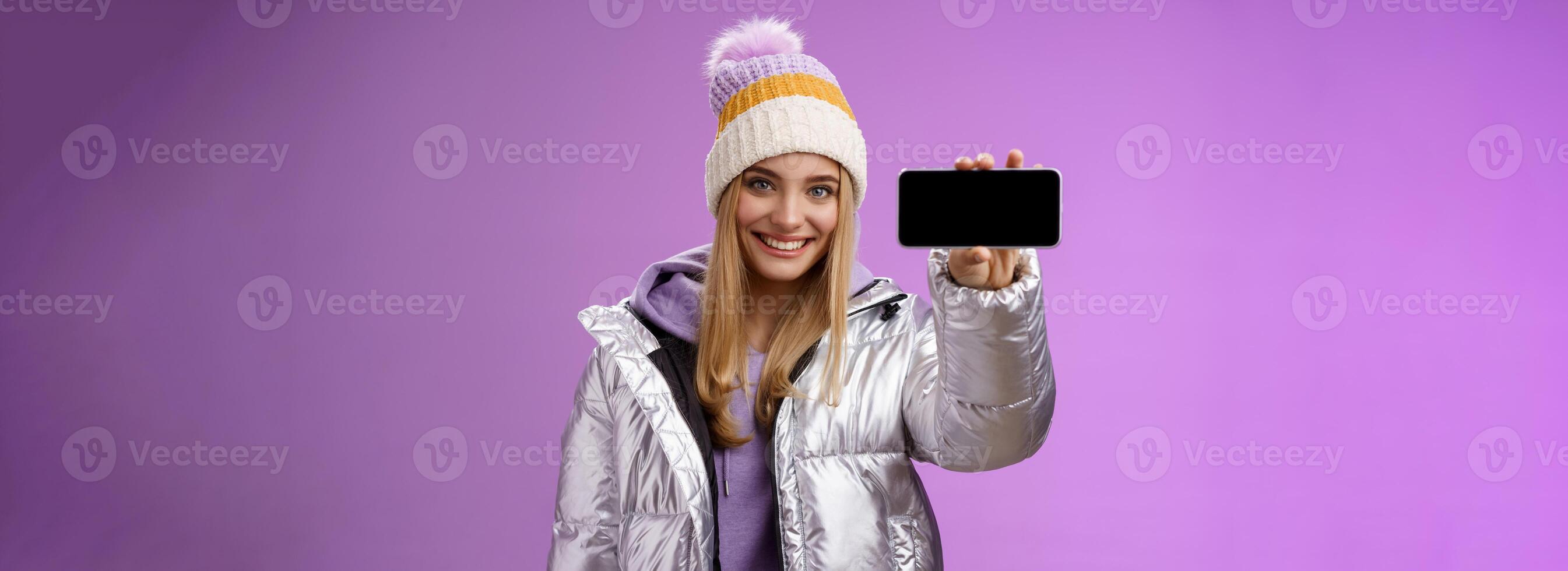 zuversichtlich freundlich gut aussehend blond Mädchen im draussen Silber glänzend Jacke Hut halt Smartphone horizontal zeigen Handy, Mobiltelefon Telefon Anzeige durchsetzungsfähig Lächeln empfehlen verwenden Anwendung, lila Hintergrund foto