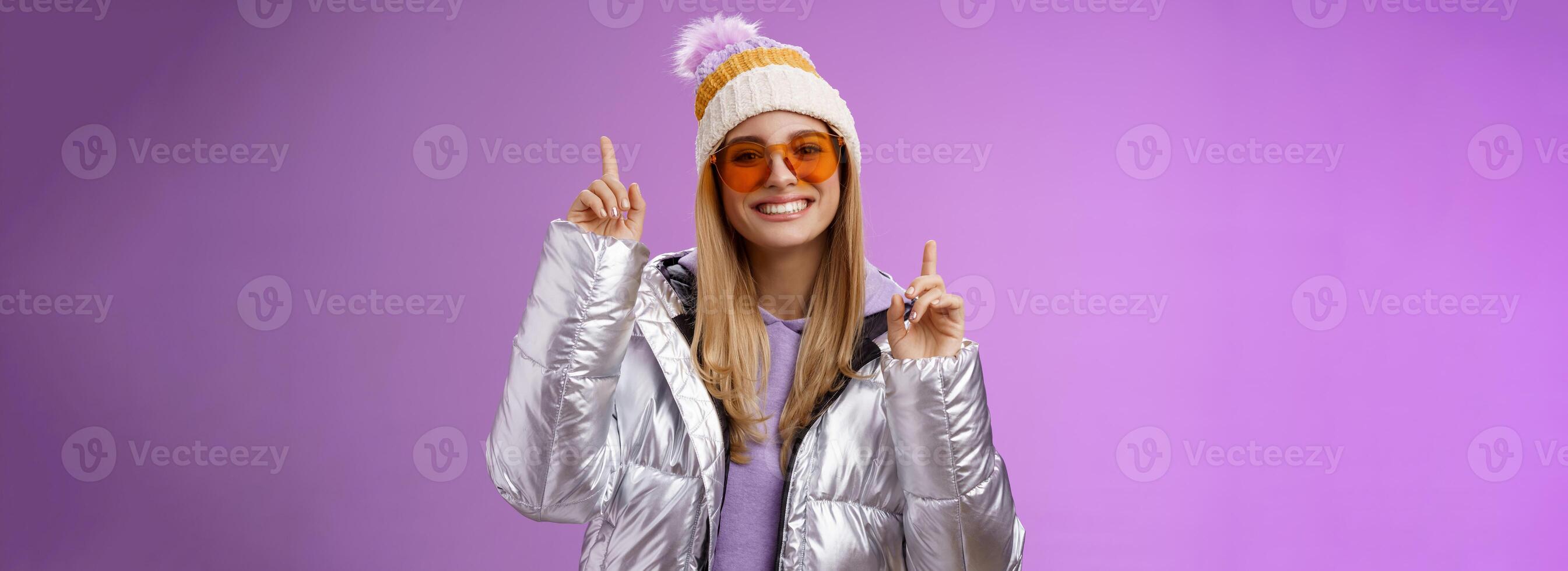froh erregt unterhalten süß blond Frau haben Spaß genießen Ferien schneebedeckt Berg Ausflug tragen Sonnenbrille Silber Jacke Winter Hut Tanzen zeigen oben amüsiert Stehen lila Hintergrund foto