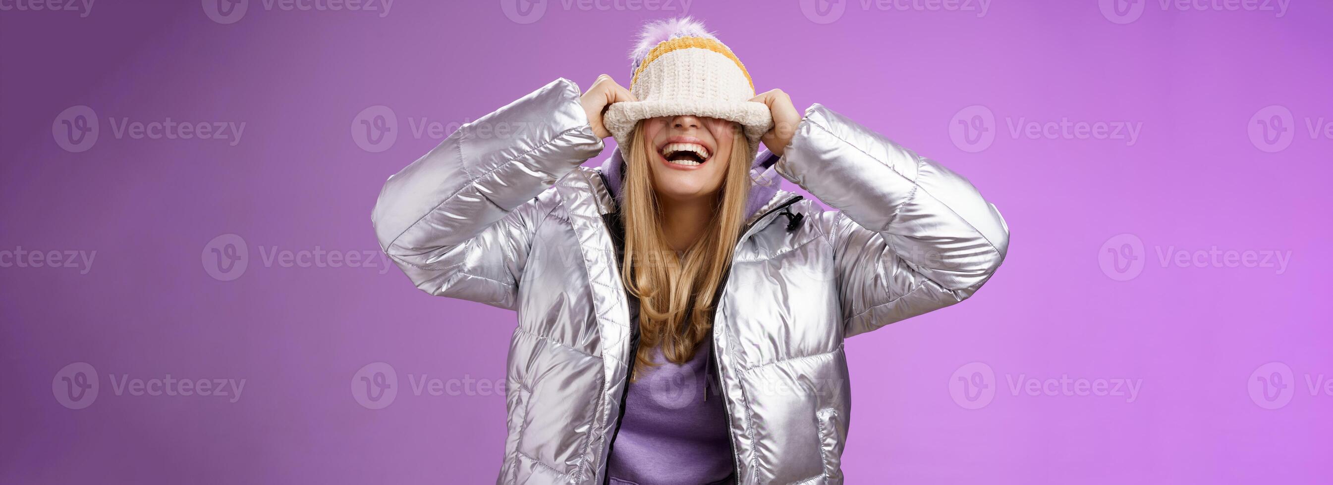 Erfreut sorglos charmant glücklich jung blond Frau haben Spaß versteckt Gesicht ziehen Hut Augen lächelnd Lachen freudig genießen Winter sonnig Tag spielen Geschwister Stehen amüsiert lila Hintergrund foto