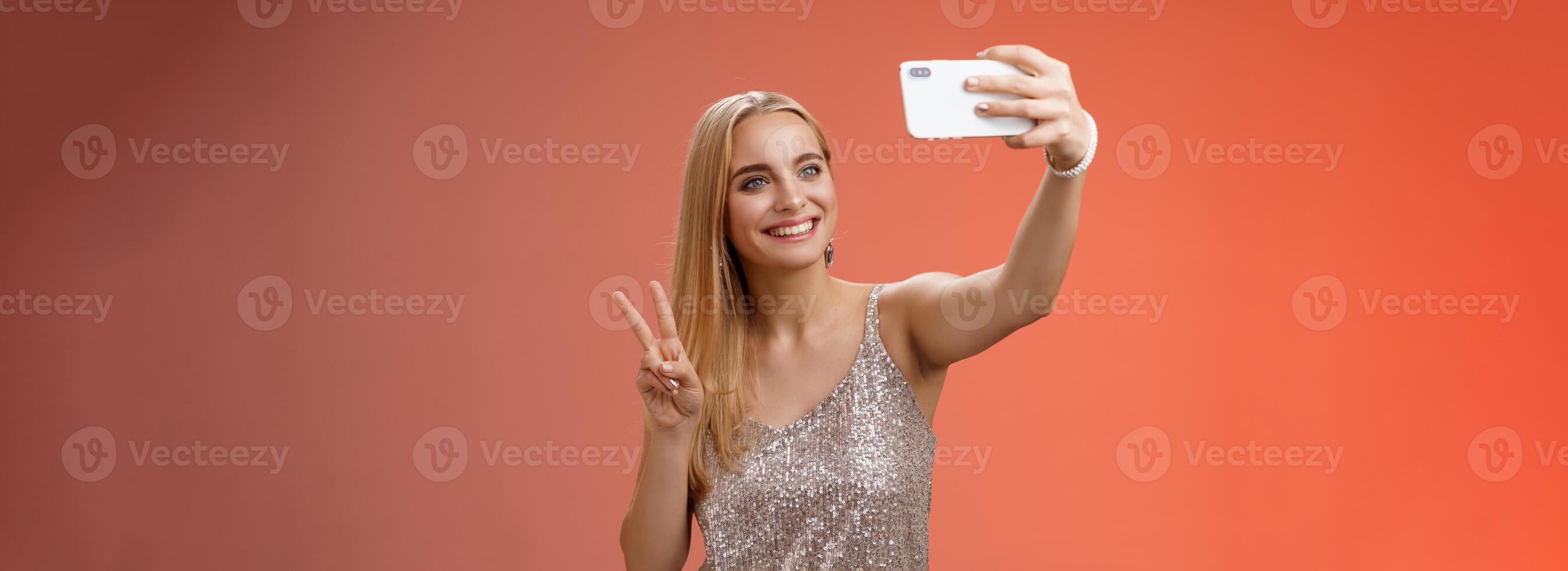 attraktiv feminin zärtlich jung blond Mädchen 25s im Silber stilvoll Kleid nehmen Selfie erweitern Arm oben Show Frieden Geste Smartphone Anzeige Aufzeichnung Video Gruß Internet Fans, Stehen rot Hintergrund foto