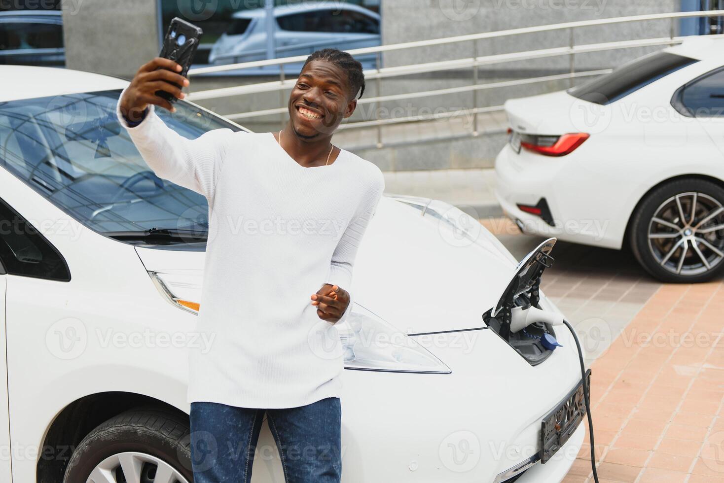 afrikanisch Mann verwenden Clever Telefon während warten und Leistung liefern verbinden zu elektrisch Fahrzeuge zum Laden das Batterie im Auto foto