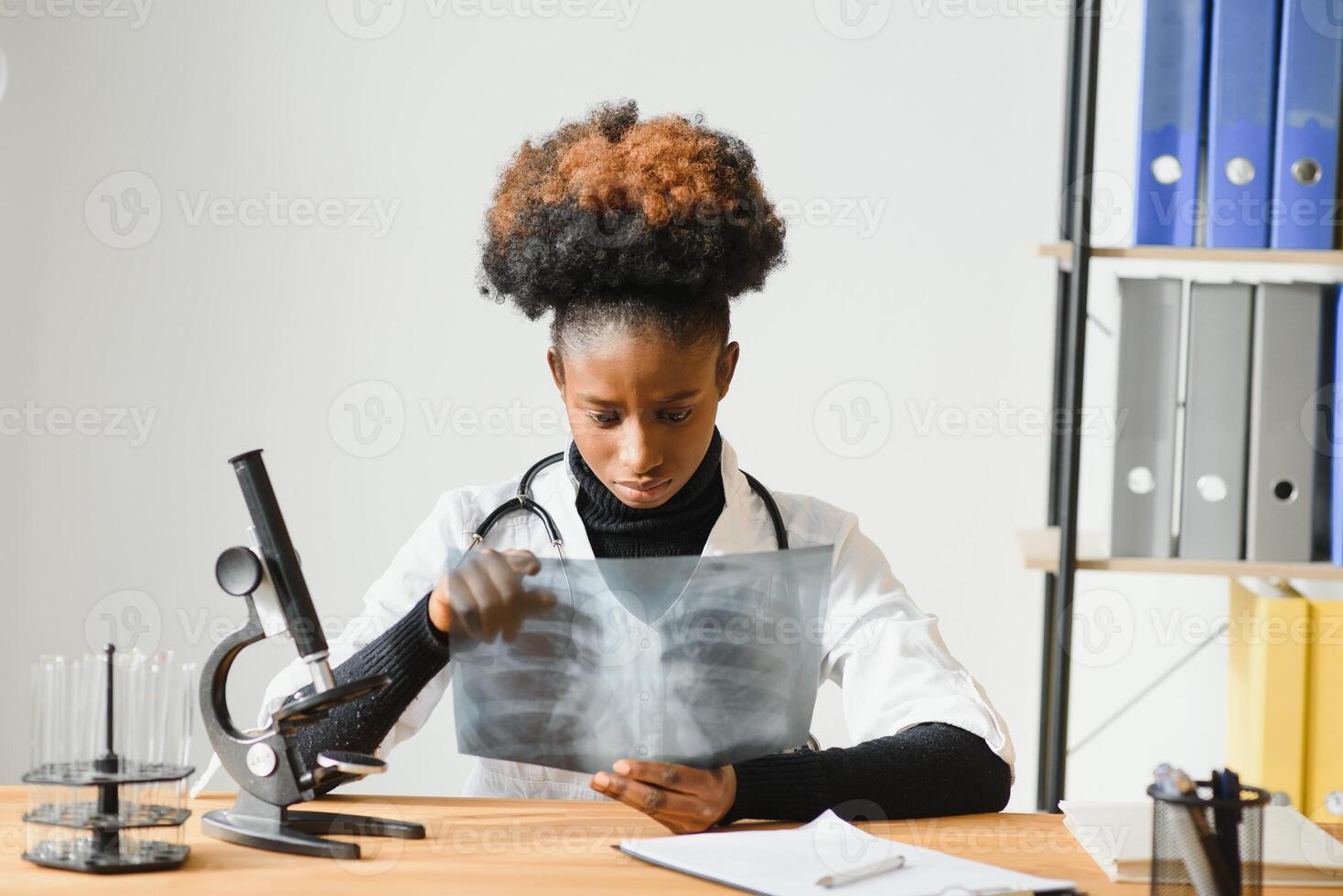 weiblich afrikanisch amerikanisch Arzt tragen Weiß Mantel mit Stethoskop Sitzung hinter Schreibtisch im das Büro und suchen Röntgen Bild. foto