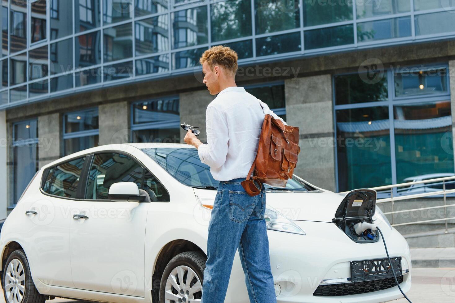 Mann Laden seine Luxus elektrisch Auto beim draussen Bahnhof im Vorderseite von modern Neu Stadt Gebäude foto