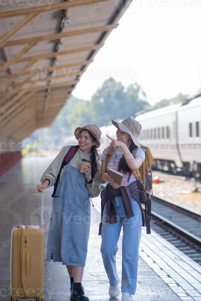 zwei jung asiatisch freunde Mädchen mit Rucksäcke beim Eisenbahn Bahnhof warten zum Zug, zwei schön Frauen Gehen entlang Plattform beim Zug Bahnhof foto