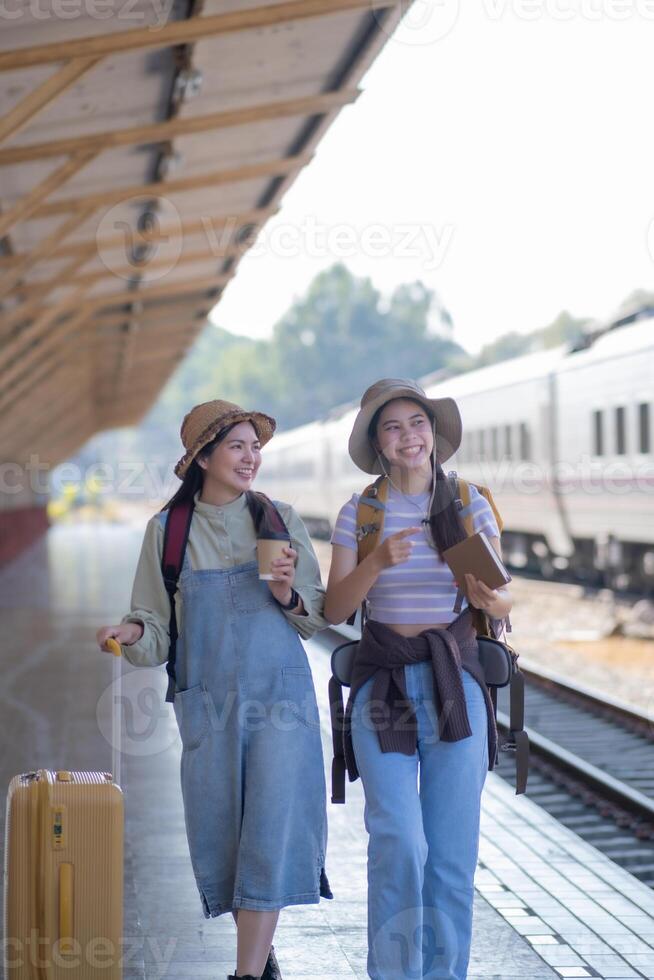 zwei jung asiatisch freunde Mädchen mit Rucksäcke beim Eisenbahn Bahnhof warten zum Zug, zwei schön Frauen Gehen entlang Plattform beim Zug Bahnhof foto