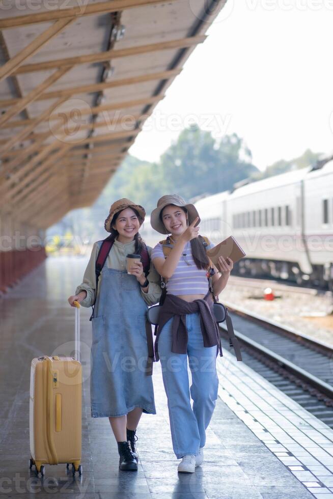 zwei jung asiatisch freunde Mädchen mit Rucksäcke beim Eisenbahn Bahnhof warten zum Zug, zwei schön Frauen Gehen entlang Plattform beim Zug Bahnhof foto