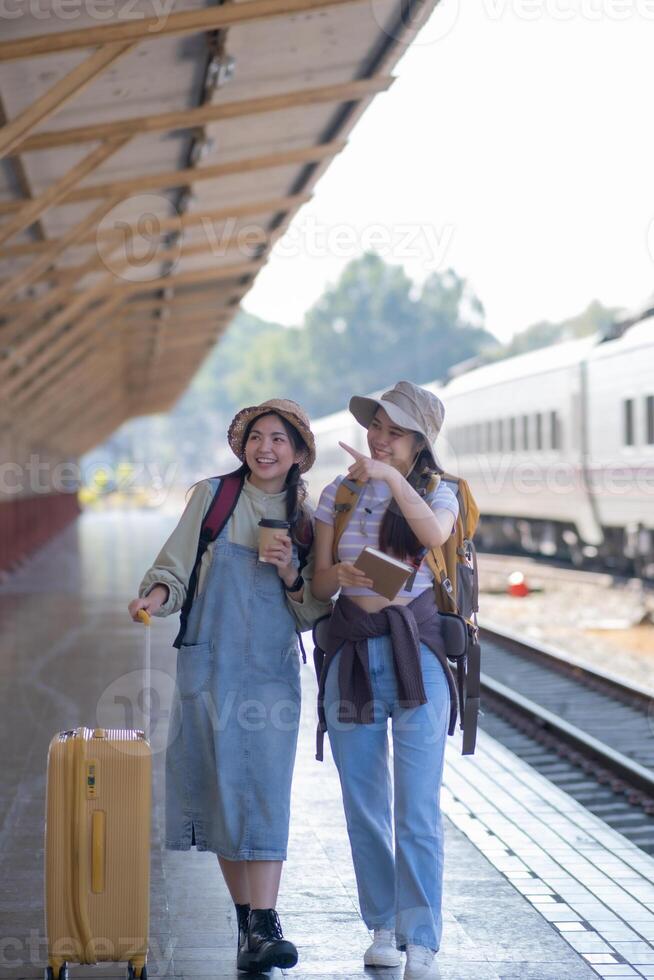 zwei jung asiatisch freunde Mädchen mit Rucksäcke beim Eisenbahn Bahnhof warten zum Zug, zwei schön Frauen Gehen entlang Plattform beim Zug Bahnhof foto