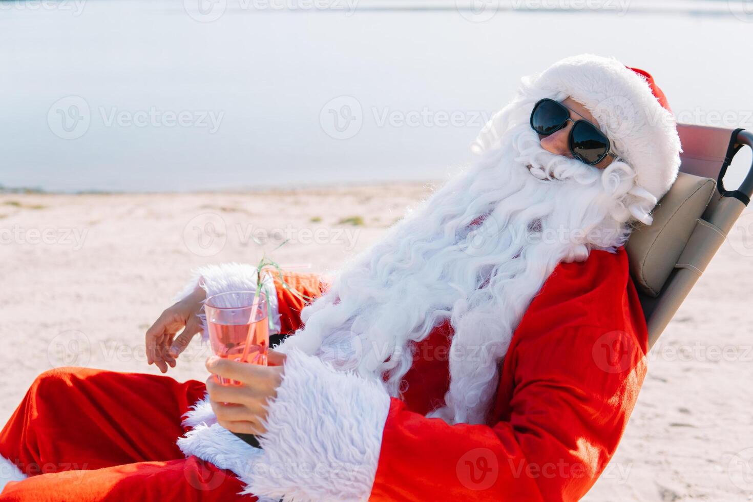 Santa claus im Sonnenbrille Getränke ein Cocktail während Lügen auf ein Sonne Liege auf das See Strand. Santa im das Tropen foto