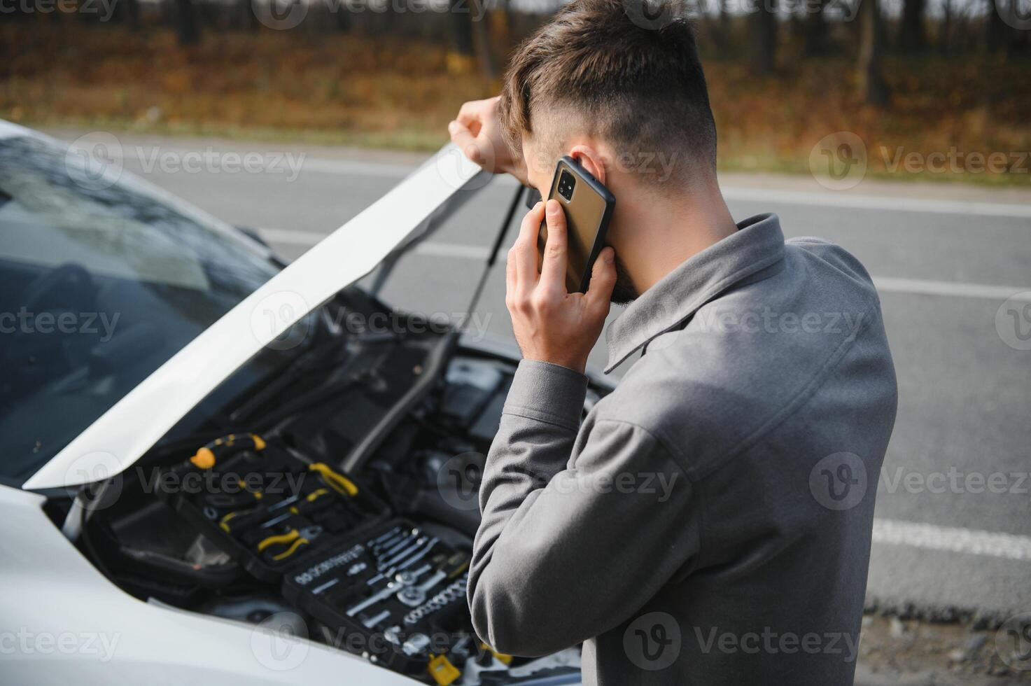 Mann verwenden ein Handy Anruf Garage im Vorderseite von das öffnen Kapuze von ein gebrochen Auto auf das Straße im das Wald. Auto Nervenzusammenbruch Konzept. foto