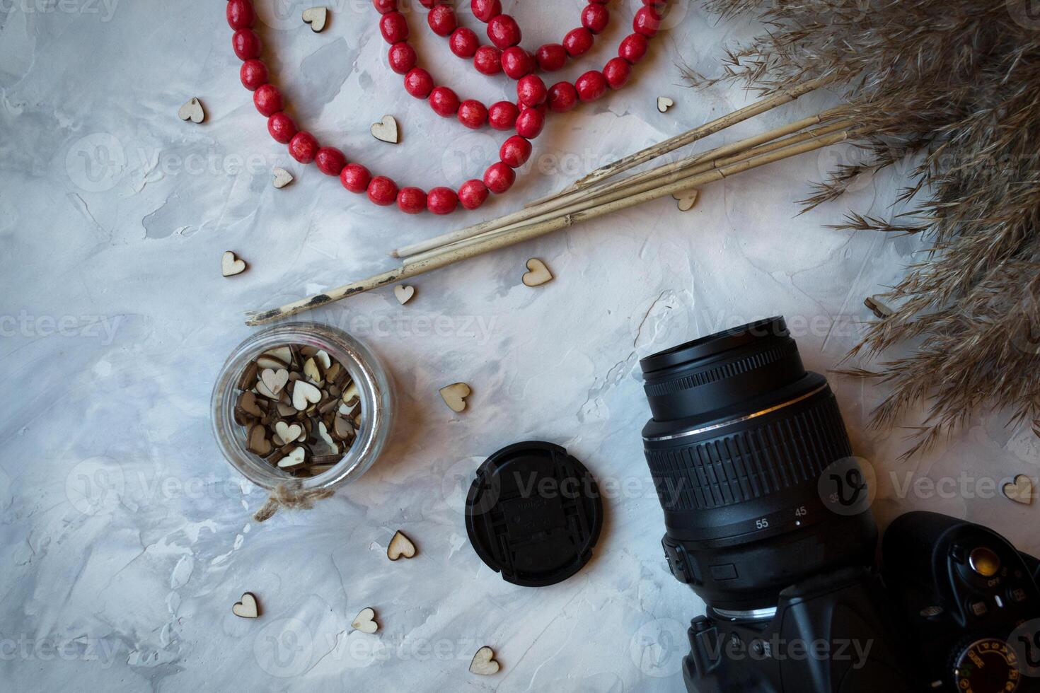 Kamera, rot Perlen, hölzern Herzen und Rohrkolben Pflanze auf ein grau texturiert Hintergrund. stilvoll immer noch Leben. foto