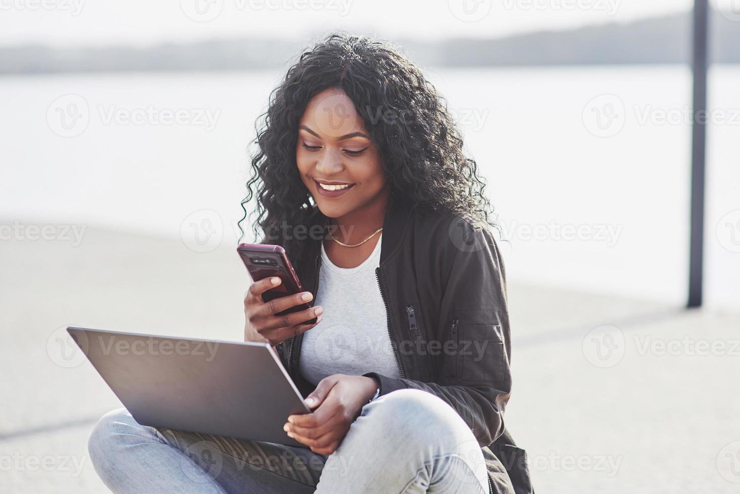 junge Frau auf der Straße, die am Laptop arbeitet und mit dem Handy spricht foto