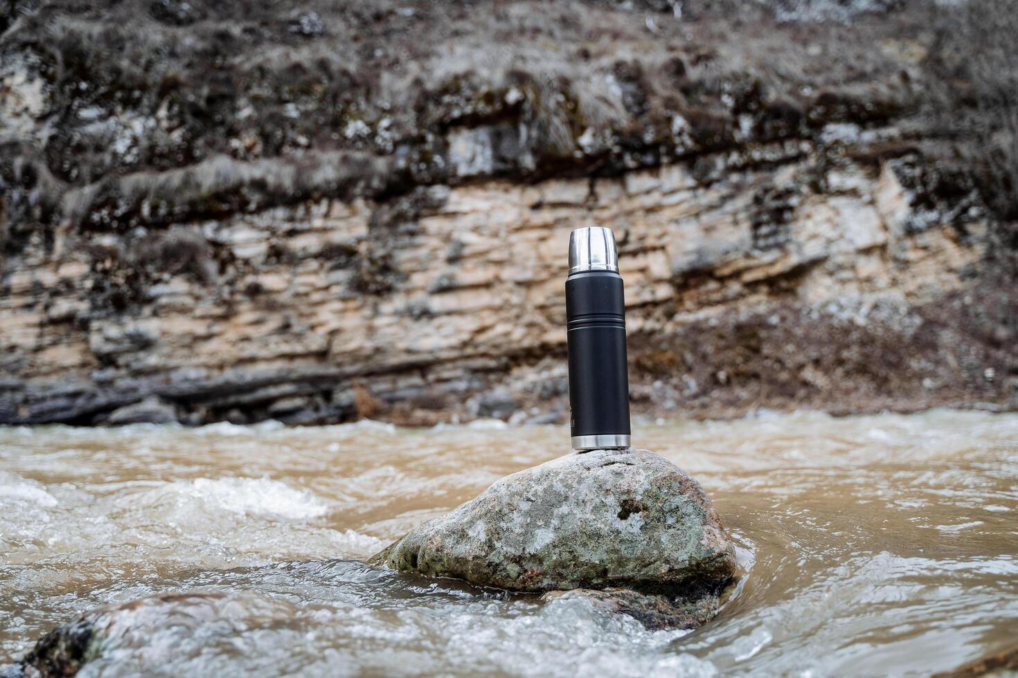 das Konzept von ein Metall Vakuum Thermosflasche gegen das Hintergrund von Natur, ein thermo Flasche steht auf ein Stein im das Fluss, Wandern Frühling Felsen. Tourist Utensilien. foto