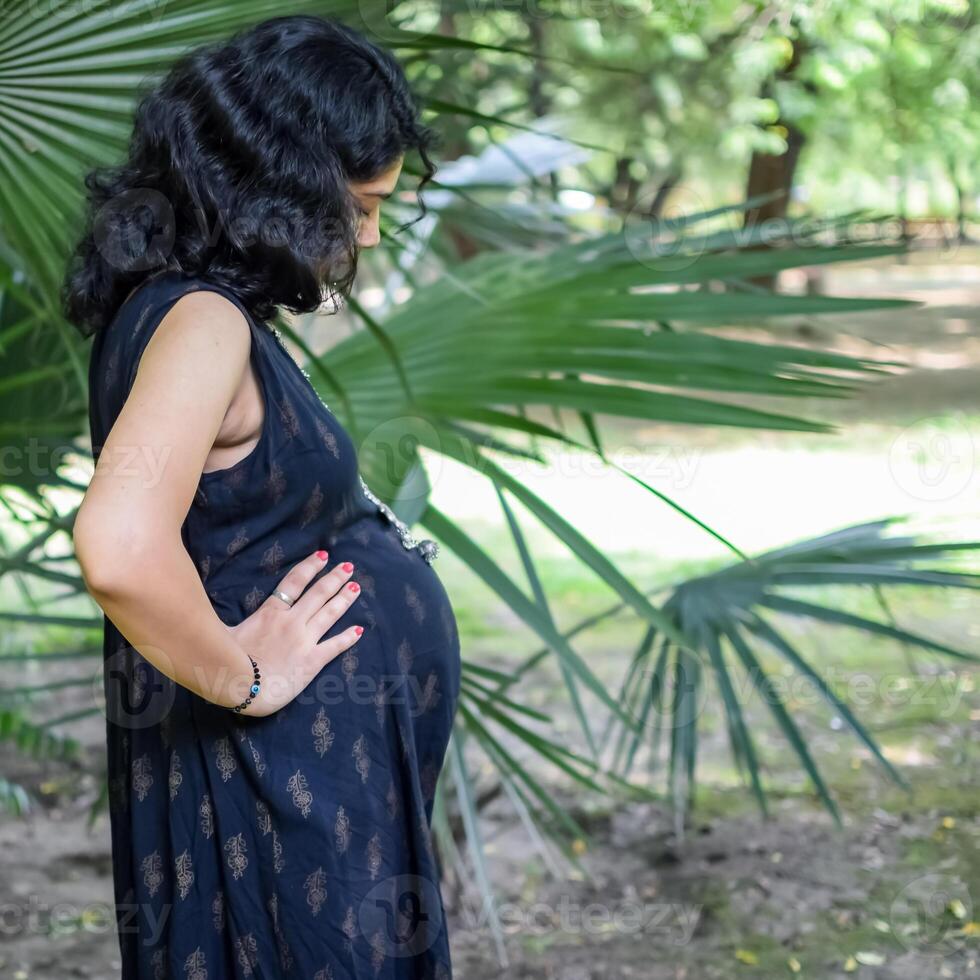 ein schwanger indisch Dame posiert zum draussen Schwangerschaft schießen und Hände auf Bauch, indisch schwanger Frau setzt ihr Hand auf ihr Bauch mit ein Mutterschaft Kleid beim Gesellschaft Park, schwanger draußen Mutterschaft schießen foto