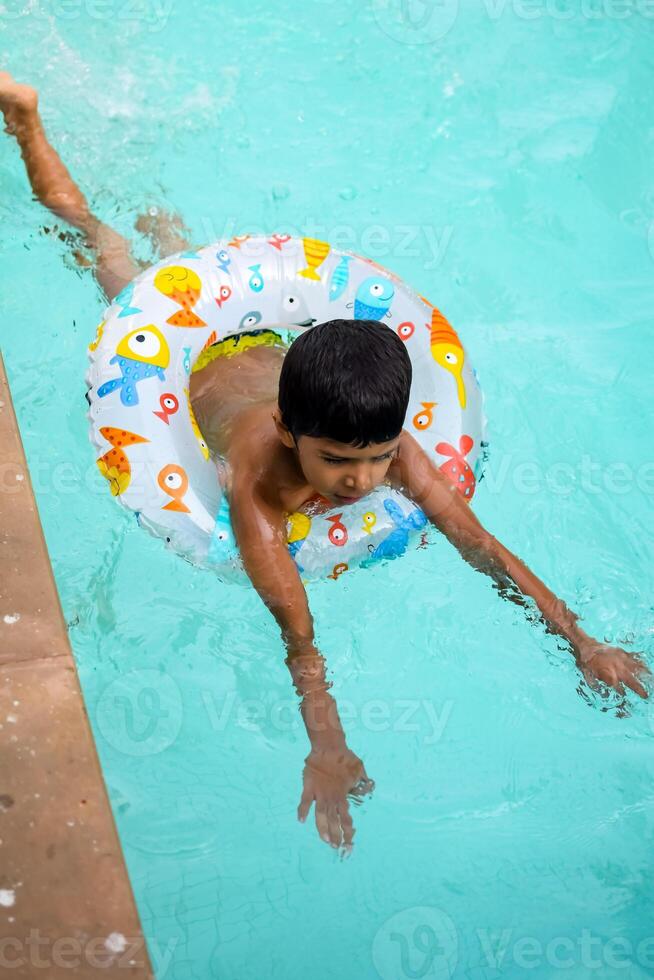 glücklich indisch Junge Schwimmen im ein Schwimmbad, Kind tragen Schwimmen Kostüm entlang mit Luft Tube während heiß Sommer- Urlaube, Kinder Junge im groß Schwimmen Schwimmbad. foto