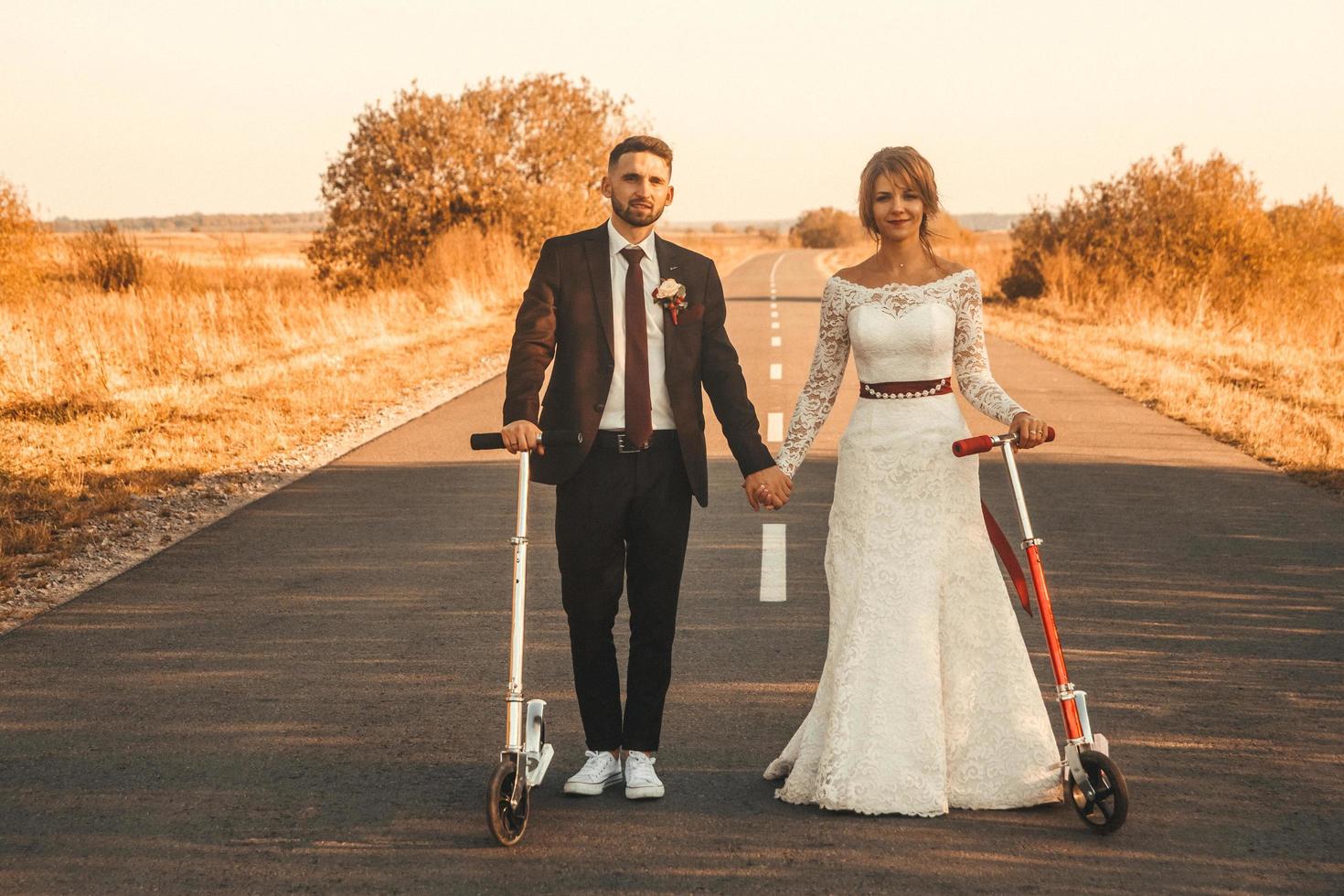 lächelndes Hochzeitspaar, das bei Sonnenuntergang auf Rollern entlang der Straße außerhalb der Stadt fährt. foto