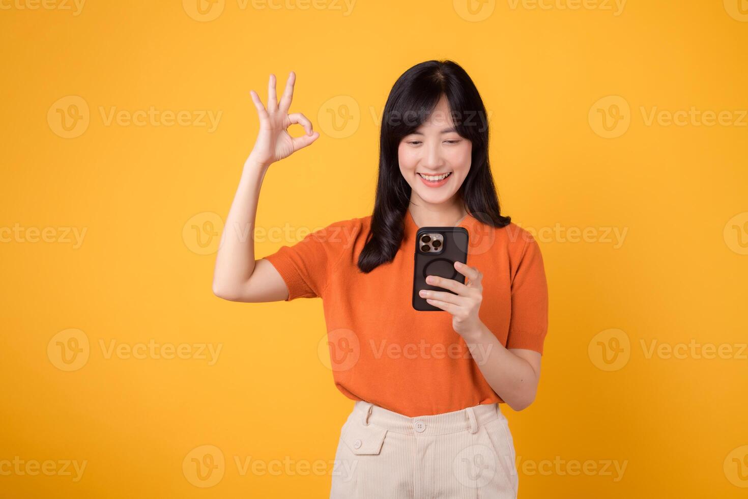energisch asiatisch Frau im ihr 30er Jahre, tragen Orange Shirt, mit Smartphone mit okay Hand Zeichen auf beschwingt Gelb Hintergrund. Neu Handy, Mobiltelefon App Konzept. foto