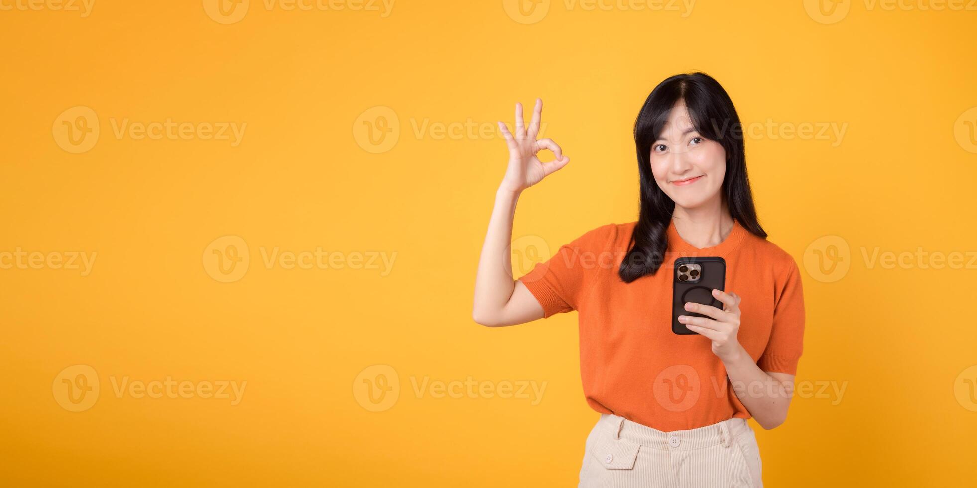 beschwingt asiatisch Frau im ihr 30er Jahre, tragen Orange Shirt, mit Smartphone mit okay Hand Zeichen auf beschwingt Gelb Hintergrund. Neu Handy, Mobiltelefon App Konzept. foto