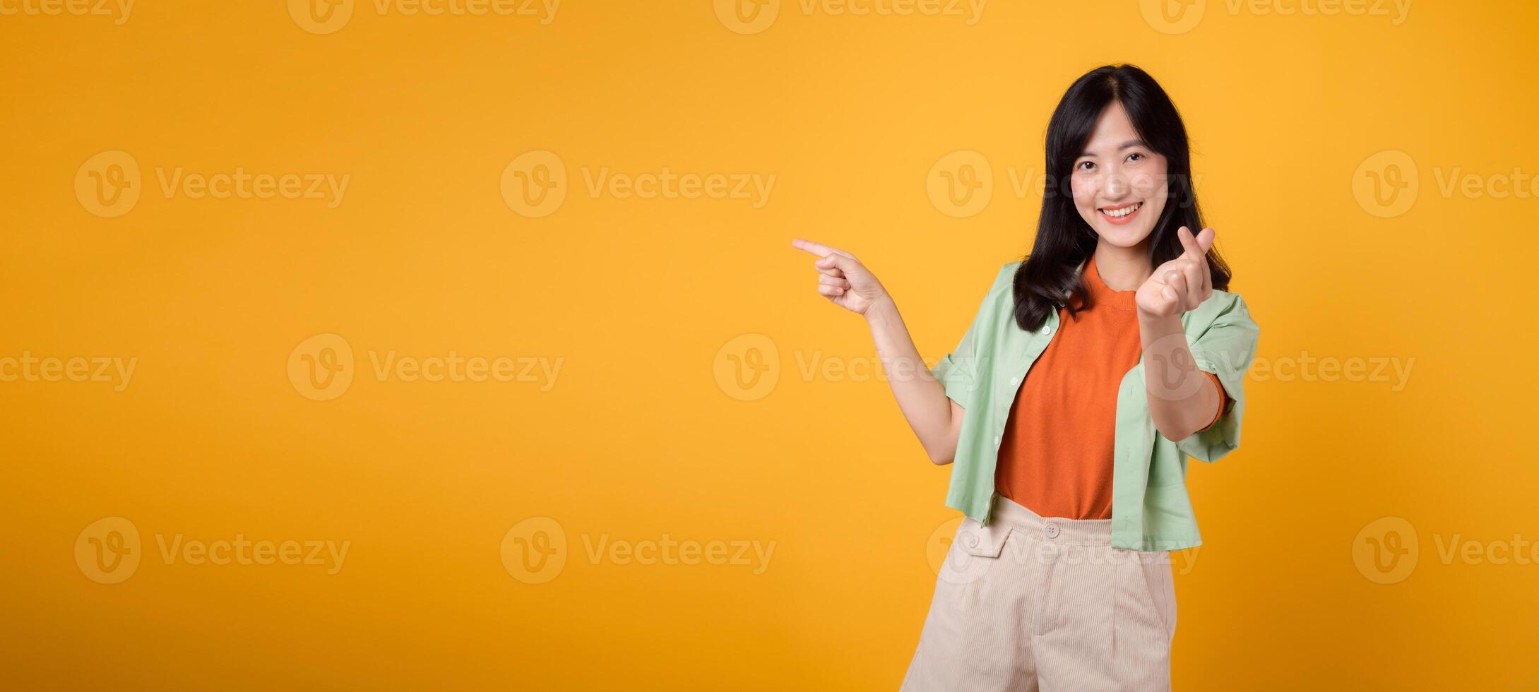 jung 30er Jahre asiatisch Frau tragen ein Grün Hemd auf ein Orange Hintergrund Punkte Finger zu kostenlos Kopieren Raum während zeigen ein Mini Herz Hand unterzeichnen. erkunden das Welt von Rabatt Einkaufen Beförderung Konzept. foto