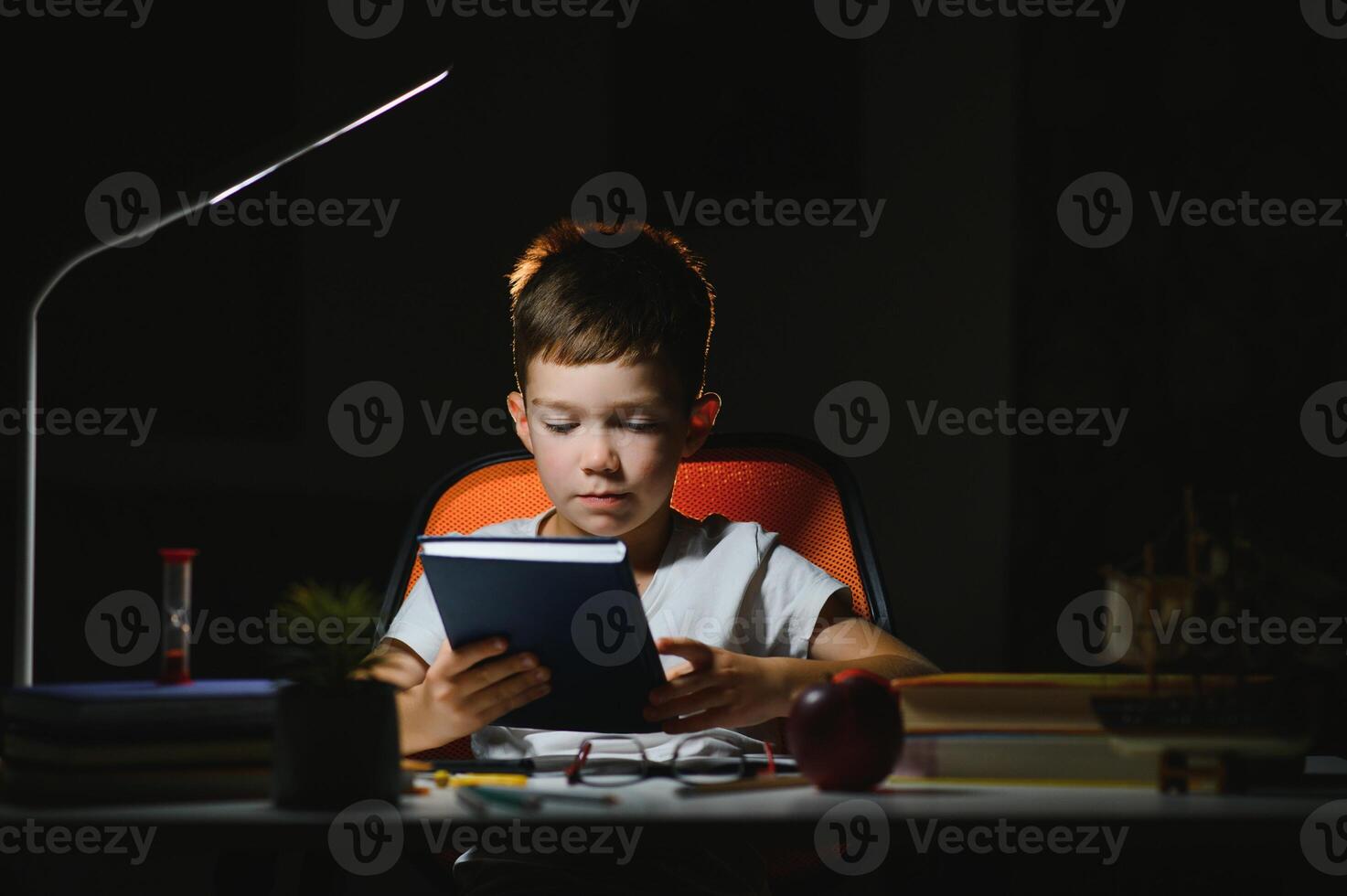 konzentriert Schüler lesen Buch beim Tabelle mit Bücher, Anlage, Lampe, Farbe Bleistifte, Apfel, und Lehrbuch foto