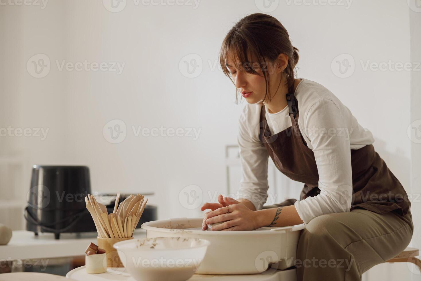 konzentriert weiblich Handwerker im Schürze Sitzung auf Bank mit Keramik Rad und Herstellung Lehm Topf foto