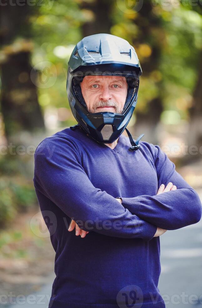 ein Senior Mann im dunkel Helm draussen. beiläufig Kleidung. gekreuzt Hände. Blau Pullover. draussen verschwommen Hintergrund. Nahaufnahme. foto