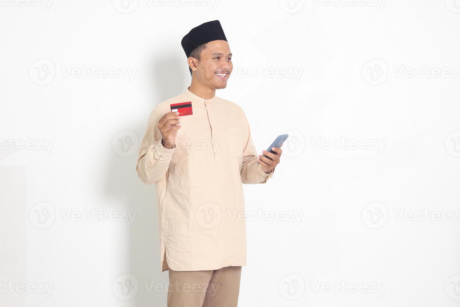 Porträt von attraktiv asiatisch Muslim Mann im koko Hemd mit Schädeldecke halten ein Handy, Mobiltelefon Telefon und präsentieren Anerkennung Karte. isoliert Bild auf Weiß Hintergrund foto