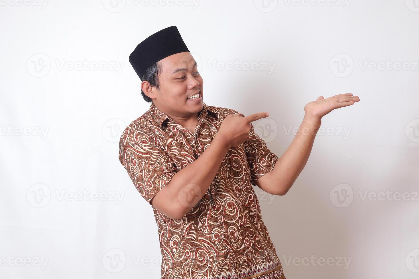 Porträt von aufgeregt asiatisch Mann tragen Batik Hemd und Songkok lächelnd und suchen beim das Kamera zeigen mit zwei Hände und Finger zu das Seite. isoliert Bild auf grau Hintergrund foto