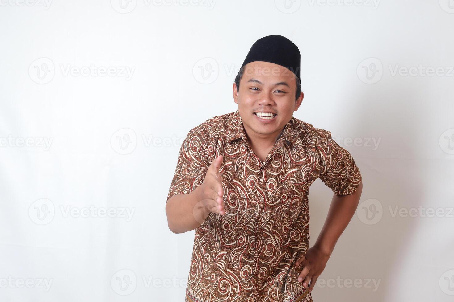 Porträt von aufgeregt asiatisch Mann tragen Batik Hemd und Songkok erreichen aus seine Hand zum ein Hand Shake, Begrüßung jemand. isoliert Bild auf grau Hintergrund foto