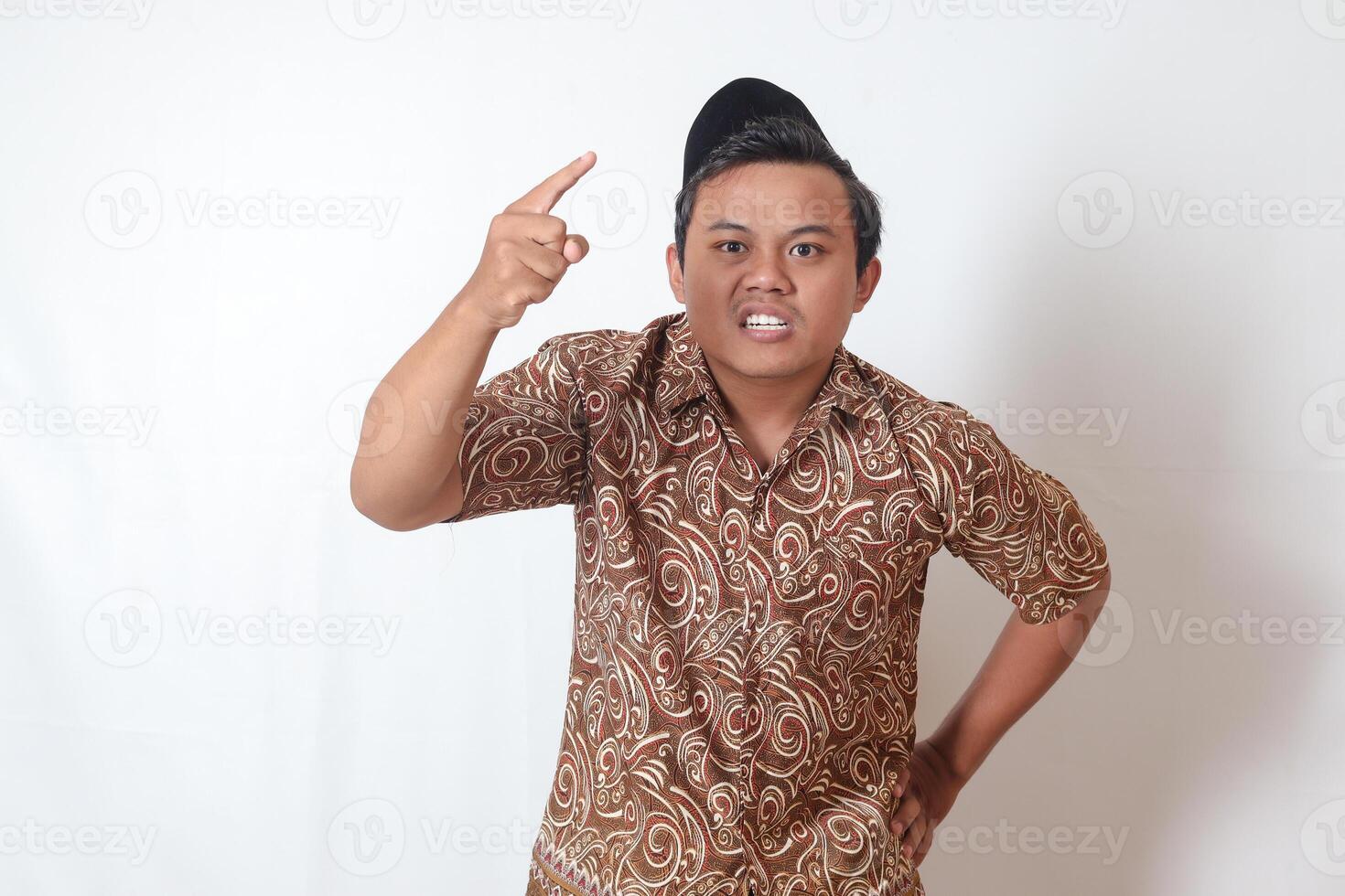 Porträt von unangenehm asiatisch Mann tragen Batik Hemd und Songkok zeigen zynisch unzufrieden wütend Gesichts- Ausdruck zeigen nach vorne, geben warnen. isoliert Bild auf grau Hintergrund foto