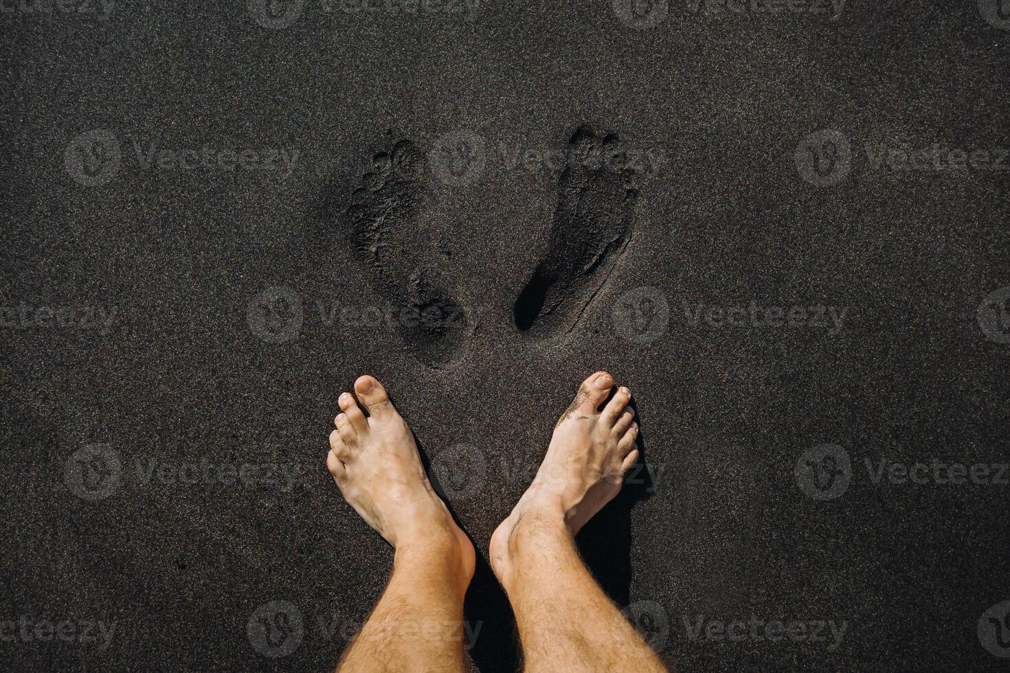 schließen oben von männlich Fußabdrücke und Füße Gehen auf das vulkanisch schwarz Sand auf das Strand foto