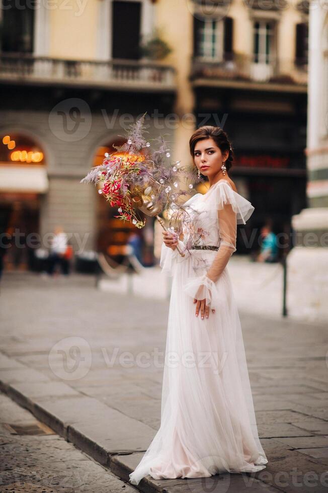 das Mädchen-Braut ist mit schön Blume Muster wie ein Maske im Florenz, stilvoll Braut im ein Hochzeit Kleid Stehen mit ein Maske im das alt Stadt, Dorf von Florenz. Modell- Mädchen im Florenz foto