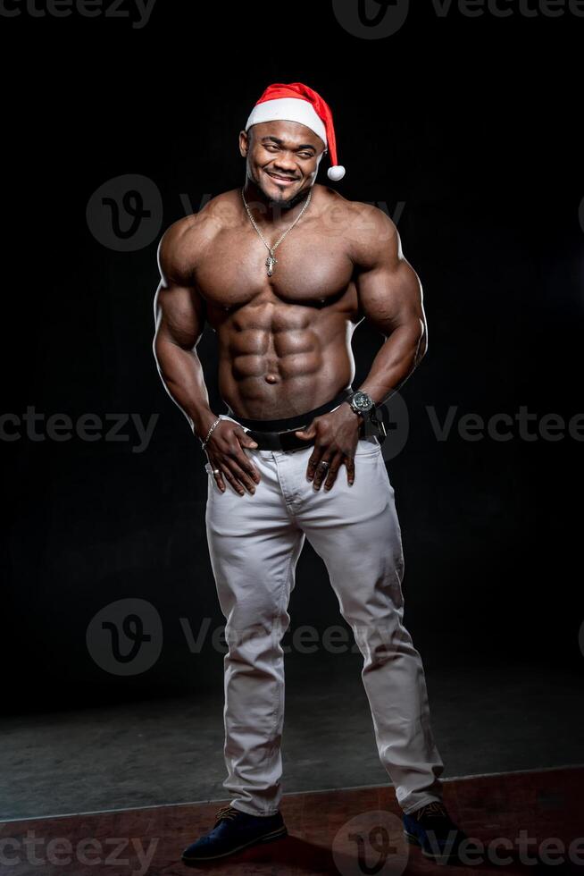 afro amerikanisch sportlich Mann im Santa Hut steht mit Hand im Tasche. sehr muskulös Körper und stark Abs. foto