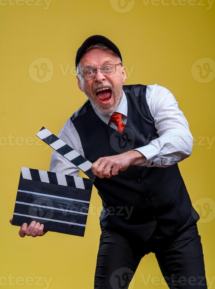 Senior gut aussehend Mann halten ein Kino Klöppel. Mann tragen passen mit Nein Jacke. Person isoliert gegen Gelb Hintergrund. Mann mit Film Klappe während filmen foto