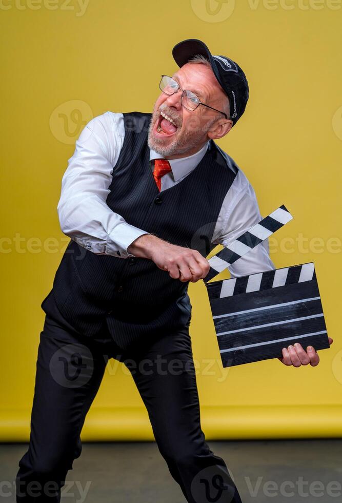 Senior gut aussehend Mann halten ein Kino Klöppel. Mann tragen passen mit Nein Jacke. Person isoliert gegen Gelb Hintergrund. foto