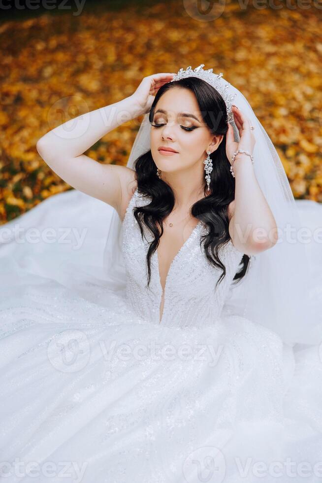 ein Brünette Braut mit ein Tiara im ihr Haar sitzt auf das Boden im ihr fließend Kleid und hält ein Strauß. auf das Hintergrund von Herbst Blätter. Hochzeit Zeremonie foto
