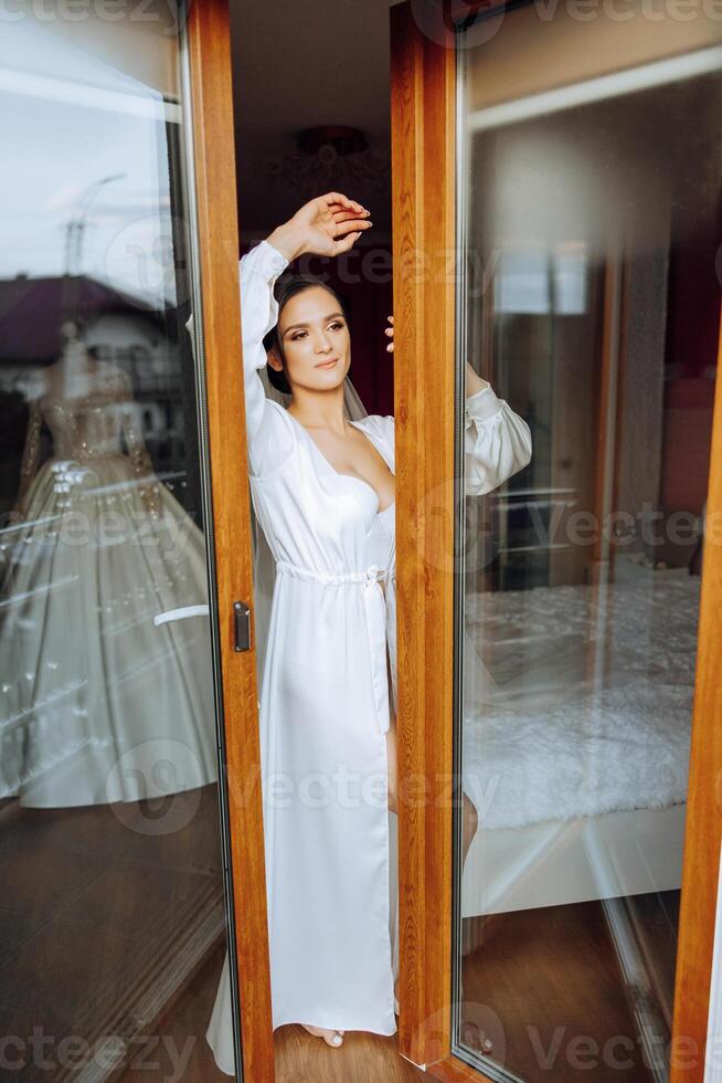 das Braut steht auf das Balkon zwischen das öffnen Fenster im ein Weiß Kleid und sieht aus voraus. Hochzeit Tag. ein Herrlich Braut. Hochzeit. foto