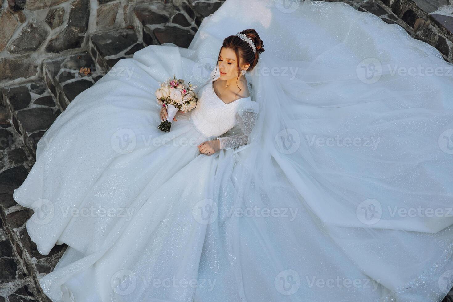 ein Brünette Braut mit ein Tiara im ihr Haar ist Lügen auf ein Kleid gelegt aus im das gestalten von ein Kreis, posieren während halten ein Strauß. auf ein Weiß Hintergrund. sonnig Tag. Hochzeit Zeremonie foto