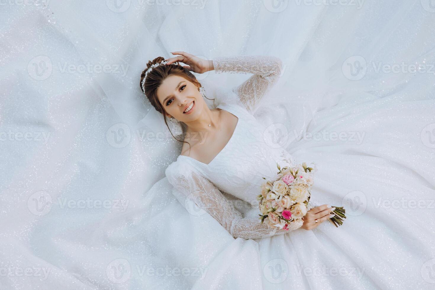 ein Brünette Braut Lügen auf ein Spitze Kleid, posiert halten ein Strauß und ihr Tiara. schön bilden und Haar. auf ein Weiß Hintergrund. sonnig Tag. Hochzeit Zeremonie foto